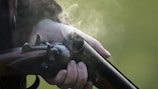 A hand tightly grips a vintage double-barrel shotgun, with smoke emanating from its recently fired barrel. The background is blurred, likely indicating an outdoor setting.