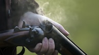 A hand tightly grips a vintage double-barrel shotgun, with smoke emanating from its recently fired barrel. The background is blurred, likely indicating an outdoor setting.