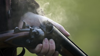 A hand tightly grips a vintage double-barrel shotgun, with smoke emanating from its recently fired barrel. The background is blurred, likely indicating an outdoor setting.