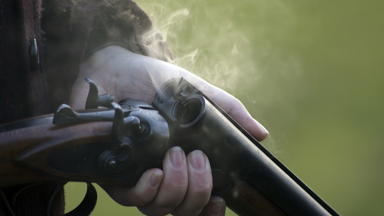 A hand tightly grips a vintage double-barrel shotgun, with smoke emanating from its recently fired barrel. The background is blurred, likely indicating an outdoor setting.