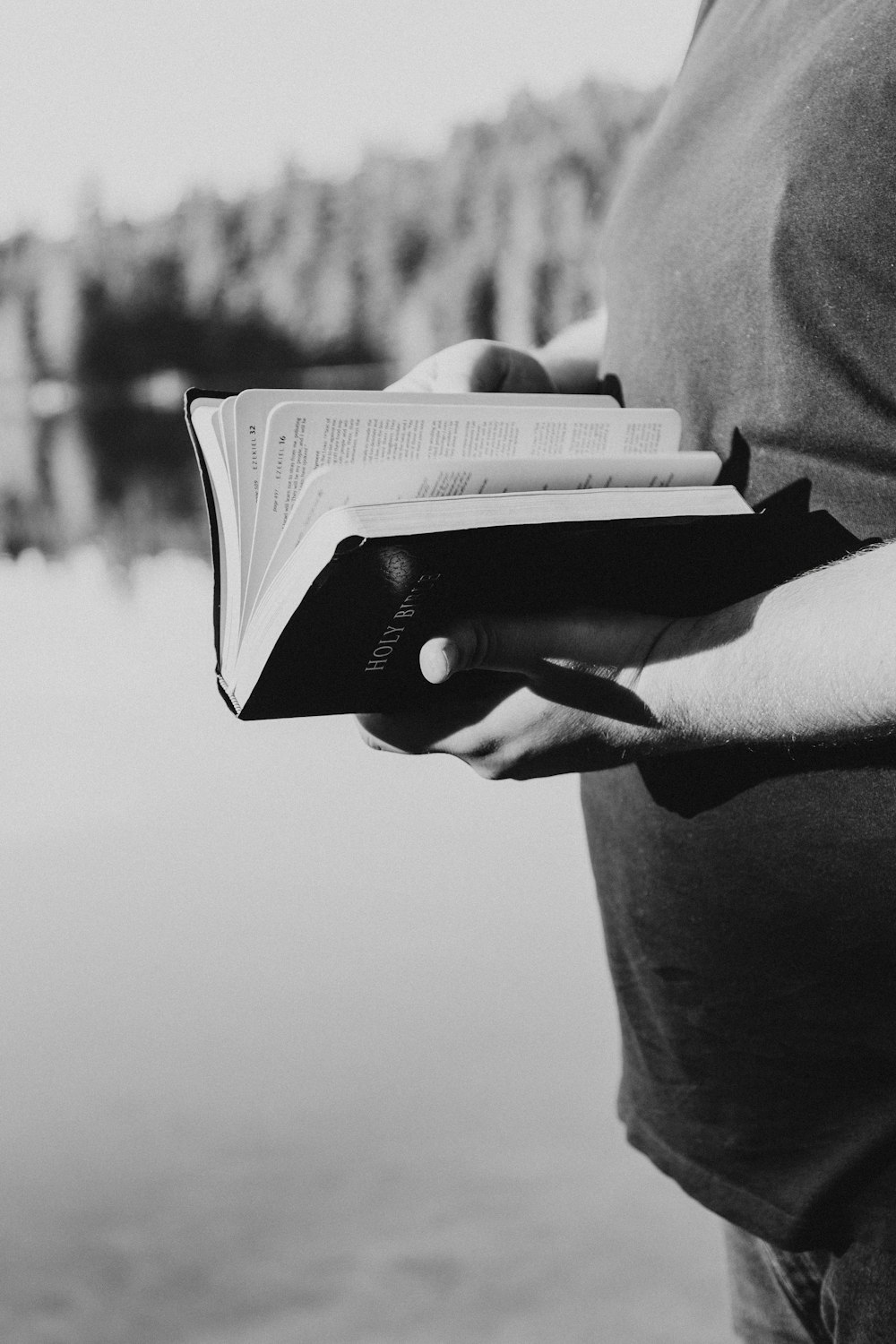 grayscale photography of person standing in front of body of water while holding book