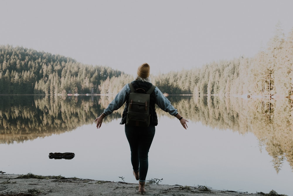 Frau in schwarzer Hose und grauem Kapuzenpullover trägt Rucksack und steht vor dem See