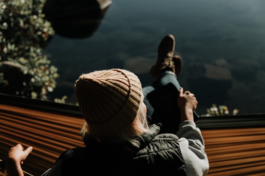 A person wearing a knitted beanie and a vest is lounging in a hammock. The setting appears tranquil, possibly near a body of water, with blurred foliage in the background. The person's legs are stretched out, and they are holding onto one end of the hammock.