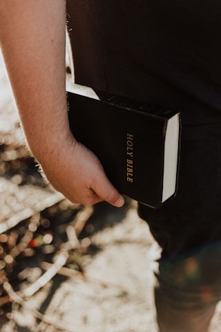 A person is holding a black book labeled 'Holy Bible' in their hand. The image captures a close-up view of the book and hand, with a blurred outdoor background.