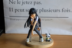 A figurine of an animated girl with long black hair sitting on a chair. She appears thoughtful with her head resting on her hand. Next to her is a small panda figure holding bamboo, both placed on a round wooden base. The background has French text printed on paper.