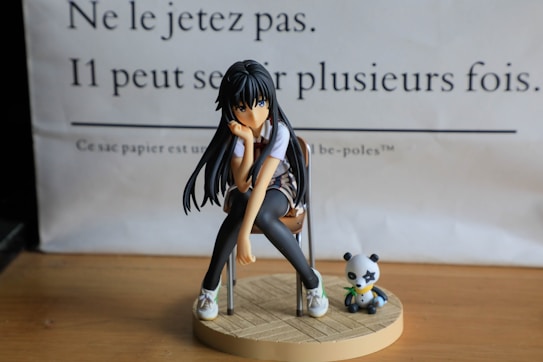A figurine of an animated girl with long black hair sitting on a chair. She appears thoughtful with her head resting on her hand. Next to her is a small panda figure holding bamboo, both placed on a round wooden base. The background has French text printed on paper.