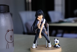 A figure of an anime-style female character with long black hair sits thoughtfully on a chair, supported by a circular base. She wears casual clothing, including a white shirt and black leggings, paired with white sneakers. Beside her, a small panda figure with a star on its face sits on the base. A water bottle with a bear illustration is partially visible on the left side of the image. The background features a softly blurred interior setting with chairs and tables in muted tones.