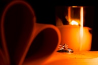 A romantic setting with a lit candle casting a warm glow. In the foreground, two rings are elegantly placed, reflecting the candlelight. To the left, pages of a book are folded into the shape of a heart, adding to the intimate atmosphere.
