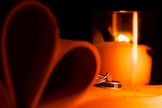 A romantic setting with a lit candle casting a warm glow. In the foreground, two rings are elegantly placed, reflecting the candlelight. To the left, pages of a book are folded into the shape of a heart, adding to the intimate atmosphere.