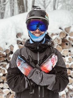 A person wearing winter gear, including a helmet with a camera and reflective goggles, stands in a snowy setting. They are smiling and holding two colorful plush fish. Stacked logs can be seen in the background.