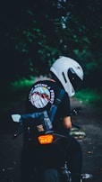 A person wearing a helmet and a backpack sits on a motorcycle, facing a forested path. The t-shirt has a colorful design on the back. The surroundings are dim, with greenery and a quiet path visible.