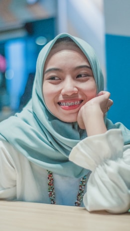 A person wearing a light blue hijab and white blouse, smiling with braces on their teeth. The background is softly blurred with shades of blue and pink.