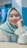 A person wearing a light blue hijab and white blouse, smiling with braces on their teeth. The background is softly blurred with shades of blue and pink.