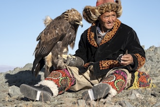 An eagle hunter in traditional attire holding a majestic golden eagle perched on his arm in a snowy landscape.