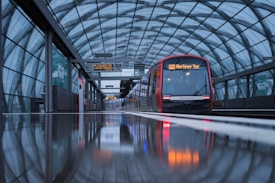 A modern, sleek train is stationed at an urban platform under an arched glass roof. The surface of the platform reflects the train and surrounding structure, suggesting a recent rainfall or high-gloss finish. Bright lights on the train's front display the destination, and there are signs and lights adding a sense of modernity and functionality.