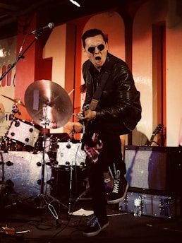 A musician in a leather jacket and sunglasses aggressively plays an electric guitar on stage, with drums and an amplifier in the background. The performer appears energetic and expressive, with one foot lifted, adding intensity to the performance.