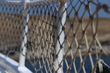 An intricately knotted rope netting attached to a white metal railing, with a blurred background of water and distant land. The netting features a series of diagonal crisscross patterns, showing wear and use.