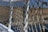 An intricately knotted rope netting attached to a white metal railing, with a blurred background of water and distant land. The netting features a series of diagonal crisscross patterns, showing wear and use.