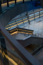 A modern architectural space featuring a multi-level staircase with sleek metal railings and illuminated steps. The surrounding walls and floors are made of smooth concrete, and large glass panels allow natural light to enter the area. The design elements emphasize geometric shapes and clean lines.