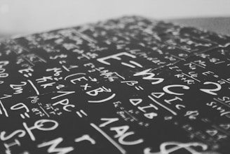 A close-up view of a chalkboard filled with various mathematical equations and symbols, written in white chalk on a dark surface. The focus is on the center, displaying formulas and expressions such as E=mc² among others.