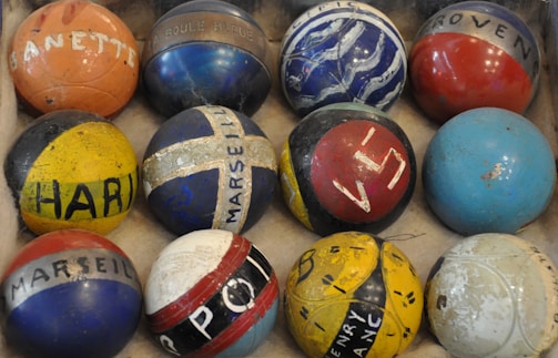 An array of colorful, vintage bowling balls with different markings and labels is arranged in a cardboard box. The balls display a variety of colors, patterns, and inscriptions, including names and numbers, adding a sense of history and personalization.