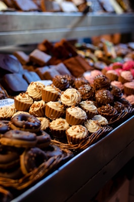 An assortment of beautifully arranged pastries and confections displayed in a bakery setting. The image features cupcakes, donuts, and various chocolates, each decorated with intricate details and toppings. The colors and textures of the sweets create a visually appealing and inviting presentation.