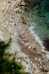 A small rocky beach with several colorful kayaks or paddleboards laid out on the sand near the water. The beach is surrounded by rugged cliffs, and the clear blue water gradually changes to darker tones further out. A few people are gathered around the equipment.