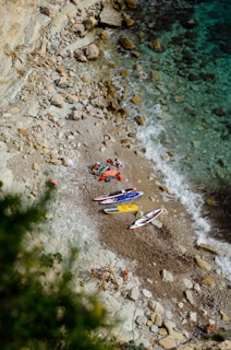 A small rocky beach with several colorful kayaks or paddleboards laid out on the sand near the water. The beach is surrounded by rugged cliffs, and the clear blue water gradually changes to darker tones further out. A few people are gathered around the equipment.