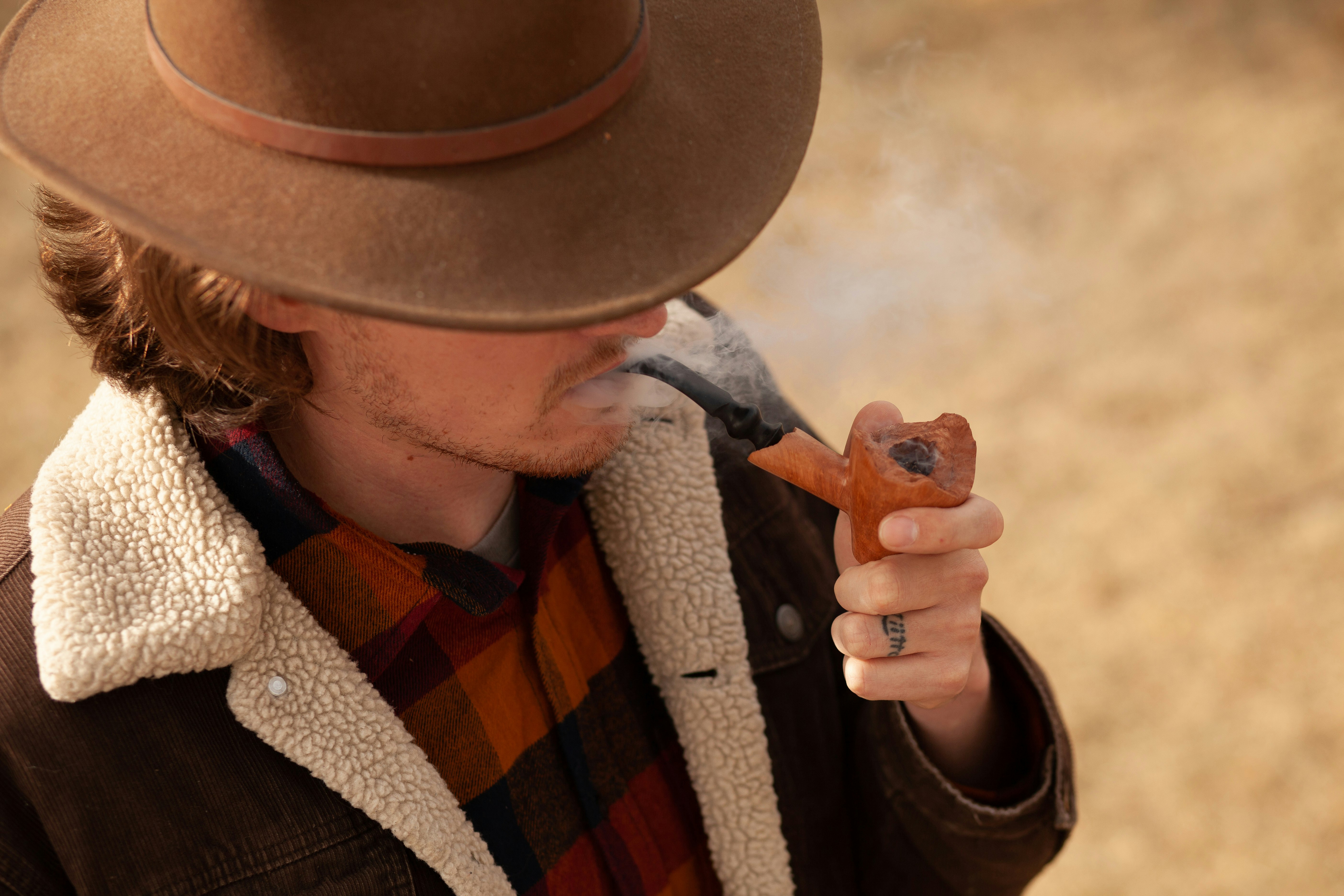 man wearing brown hat and jacket smoking tobacco pipe