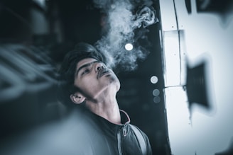 A person is leaning back, exhaling smoke, which creates a dreamy and contemplative atmosphere. The setting appears urban with bokeh lights in the background, and a strong contrast between darkness and bright light.