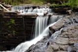 Water cascades delicately over a small wooden dam, flowing down into a rocky area below. The scene is surrounded by lush greenery, with branches and logs adding a natural element to the structure.