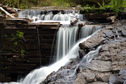 Water cascades delicately over a small wooden dam, flowing down into a rocky area below. The scene is surrounded by lush greenery, with branches and logs adding a natural element to the structure.
