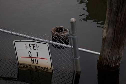 A partially submerged chain-link fence with a damaged and submerged warning sign. The water level appears high, covering the bottom part of the sign. There is a wooden post next to the fence, indicating that the area might be part of a dock or similar waterside structure. The water looks calm and slightly murky.