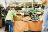 A bustling farmer's market stall features an array of fresh produce such as leafy greens, carrots, and squash neatly arranged in woven baskets. A person in a green jacket actively takes photographs of the display, while another person observes the scene. In the background, more produce is visible with handwritten price tags.