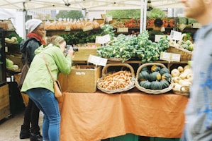 A bustling farmer's market stall features an array of fresh produce such as leafy greens, carrots, and squash neatly arranged in woven baskets. A person in a green jacket actively takes photographs of the display, while another person observes the scene. In the background, more produce is visible with handwritten price tags.