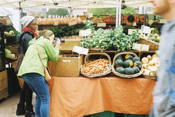 A bustling farmer's market stall features an array of fresh produce such as leafy greens, carrots, and squash neatly arranged in woven baskets. A person in a green jacket actively takes photographs of the display, while another person observes the scene. In the background, more produce is visible with handwritten price tags.
