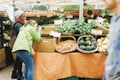 A bustling farmer's market stall features an array of fresh produce such as leafy greens, carrots, and squash neatly arranged in woven baskets. A person in a green jacket actively takes photographs of the display, while another person observes the scene. In the background, more produce is visible with handwritten price tags.