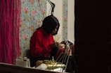 A person wearing a red sweater is carefully applying makeup to another individual's eyebrows. They are in a room with a vibrant floral wallpaper and red curtains, creating a cozy atmosphere. Various makeup tools and accessories are placed nearby.