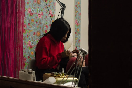 A person wearing a red sweater is carefully applying makeup to another individual's eyebrows. They are in a room with a vibrant floral wallpaper and red curtains, creating a cozy atmosphere. Various makeup tools and accessories are placed nearby.