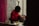 A person wearing a red sweater is carefully applying makeup to another individual's eyebrows. They are in a room with a vibrant floral wallpaper and red curtains, creating a cozy atmosphere. Various makeup tools and accessories are placed nearby.