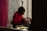 A person wearing a red sweater is carefully applying makeup to another individual's eyebrows. They are in a room with a vibrant floral wallpaper and red curtains, creating a cozy atmosphere. Various makeup tools and accessories are placed nearby.