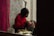 A person wearing a red sweater is carefully applying makeup to another individual's eyebrows. They are in a room with a vibrant floral wallpaper and red curtains, creating a cozy atmosphere. Various makeup tools and accessories are placed nearby.