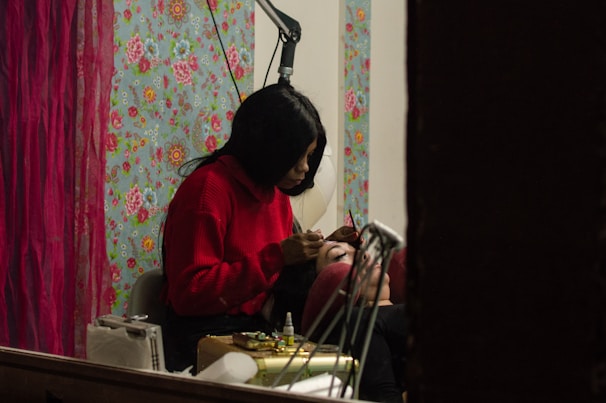 A person wearing a red sweater is carefully applying makeup to another individual's eyebrows. They are in a room with a vibrant floral wallpaper and red curtains, creating a cozy atmosphere. Various makeup tools and accessories are placed nearby.