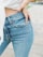 A person wearing light blue denim jeans with one hand resting on their hip. The jeans have a classic five-pocket design and appear to be high-waisted. Part of a white top and a wrist with a watch or bracelet are also visible.