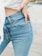 A person wearing light blue denim jeans with one hand resting on their hip. The jeans have a classic five-pocket design and appear to be high-waisted. Part of a white top and a wrist with a watch or bracelet are also visible.