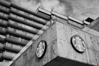 A large concrete building with multiple stories features a prominently displayed circular logo with a mermaid-like figure. The architecture has a modern, angular design. The sky appears cloudy in the background, and the image is in black and white.