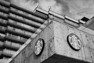 A large concrete building with multiple stories features a prominently displayed circular logo with a mermaid-like figure. The architecture has a modern, angular design. The sky appears cloudy in the background, and the image is in black and white.