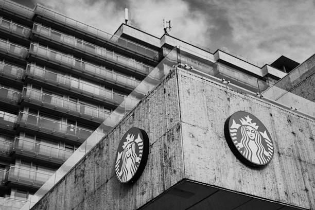 A large concrete building with multiple stories features a prominently displayed circular logo with a mermaid-like figure. The architecture has a modern, angular design. The sky appears cloudy in the background, and the image is in black and white.