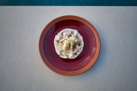 A red ceramic plate holds a gourmet dessert topped with white cream and garnished with delicate flakes and a dusting of cocoa or spices. The dish is centered on a light beige surface with a blurred blue background.