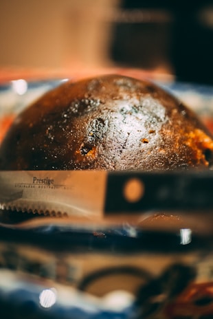 A close-up of a round, dark brown loaf of bread with a shiny crust, placed on a colorful blurred background. In the foreground, part of a serrated knife with the word 'Prestige' on it is visible.